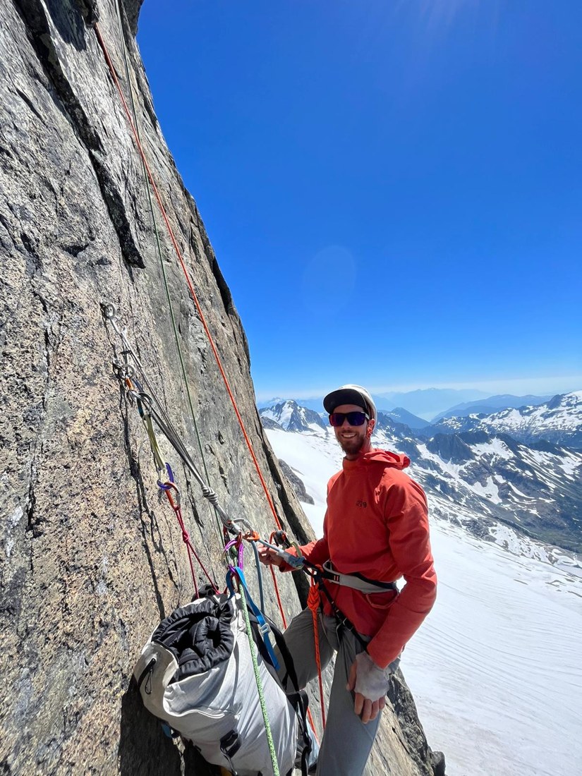 Gabriel with climbing equipment on a rock face above snow-covered mountains